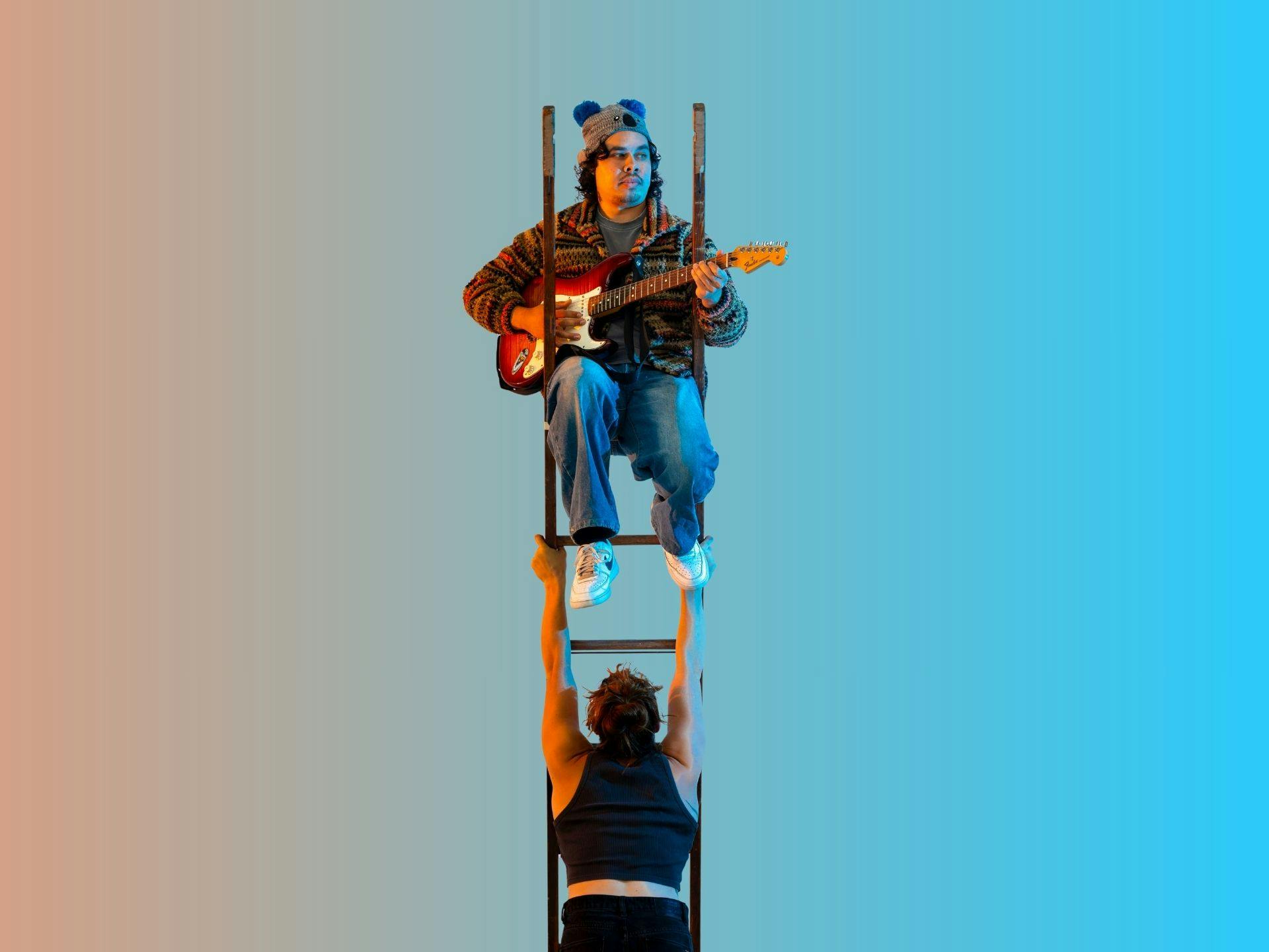 A musician sits at the top of a ladder playing a guitar. The ladder is being held upright in the air by an acrobat below. The acrobat is facing away. Photography by Tiffany Garvie